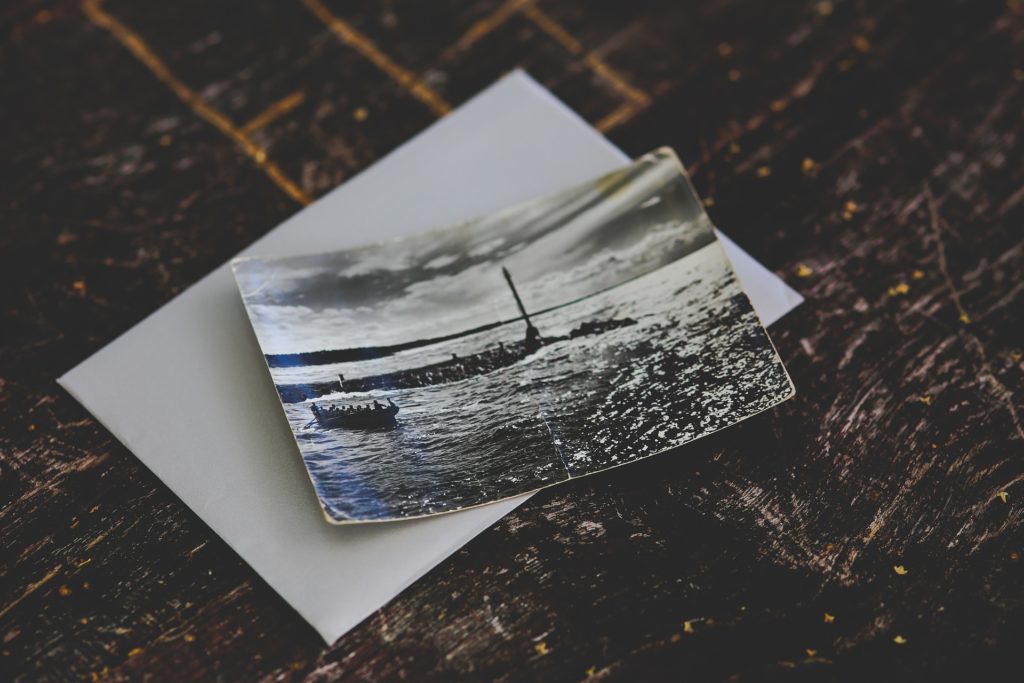 Ein altes Schwarzweißfoto liegt auf einem strukturierten, dunklen Holztisch und fängt eine ruhige Meereslandschaft mit einem Boot ein. Leicht gewellt liegt dieses Artefakt der Postkartengeschichte auf einem weißen Umschlag und erzählt Geschichten vergangener Reisen.