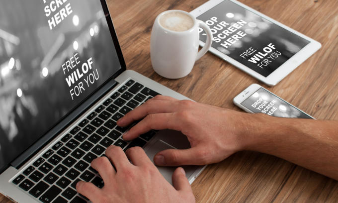 Eine Person tippt auf einem Laptop, daneben stehen ein Smartphone und ein Tablet. Auf allen Bildschirmen ist eine verschwommene Schriftwirkung zu sehen, darunter Nachrichten wie „Kostenloses WLAN für Sie“ und „Bildschirm hier löschen“. Eine weiße Tasse mit Schaum steht daneben auf einem Holztisch.