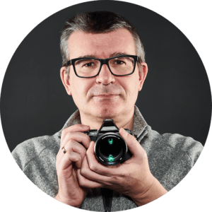 Ein Mann mit Brille und grauem Pullover steht mit einer Kamera in der Hand vor einem dunklen Hintergrund und blickt mit neutralem Gesichtsausdruck nach vorne. Dies erinnert an den klassischen Fotografiestil, der in „Das ABC der Farbe“ vorgestellt wird.