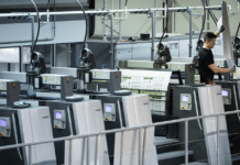 Ein Arbeiter bedient eine große, moderne Offsetdruckmaschine mit mehreren Druckwerken. Die Maschinerie umfasst Bedienfelder und Förderbänder, die die bedruckten Blätter transportieren. Diese saubere, gut beleuchtete Umgebung bietet einen industriellen Einblick in die moderne Drucktechnologie.