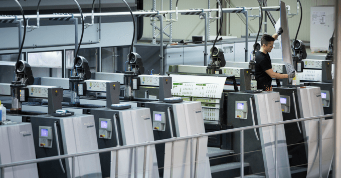 Ein Arbeiter bedient eine große, moderne Offsetdruckmaschine mit mehreren Druckwerken. Die Maschinerie umfasst Bedienfelder und Förderbänder, die die bedruckten Blätter transportieren. Diese saubere, gut beleuchtete Umgebung bietet einen industriellen Einblick in die moderne Drucktechnologie.