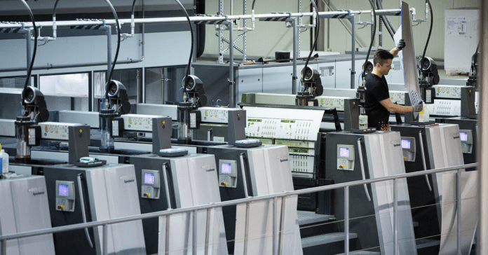 Ein Arbeiter bedient eine große, moderne Offsetdruckmaschine mit mehreren Druckwerken. Die Maschinerie umfasst Bedienfelder und Förderbänder, die die bedruckten Blätter transportieren. Diese saubere, gut beleuchtete Umgebung bietet einen industriellen Einblick in die moderne Drucktechnologie.