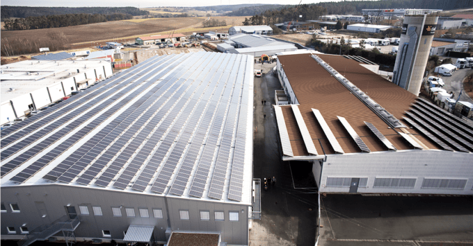 Luftaufnahme von Industriegebäuden mit Solarmodulen auf den Dächern während eines Offsetdruck-Werksrundgangs. Der Komplex ist von Feldern und anderen Industrieanlagen umgeben. Rechts steht ein Silo unter klarem Himmel.