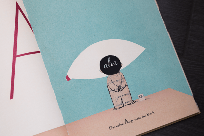 Illustration eines Kindes mit einer großen augenförmigen Sprechblase auf blauem Hintergrund. In der Sprechblase steht das Wort „aha“. Darunter steht auf Deutsch: „Das offene Auge sieht ins Buch.“ Auf der linken Seite befindet sich ein großes „A“.