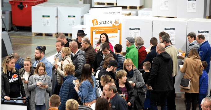 Eine große Gruppe versammelt sich an einem überdachten Bahnhof und vertieft sich in Broschüren neben einem Schild mit leuchtend orangefarbener Schrift. Es herrscht reges Treiben – möglicherweise findet eine Veranstaltung oder Ausstellung statt, die die Kunst des Offsetdruck-Werksrundgangs präsentiert und Innovationen im Druck- und Technologiebereich vorstellt.