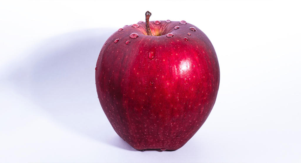 Ein frischer roter Apfel mit Wassertropfen auf der Oberfläche vor weißem Hintergrund. Der Apfel wirft einen weichen Schatten nach links, der seine glatte, glänzende Schale und seine leuchtende Farbe hervorhebt.