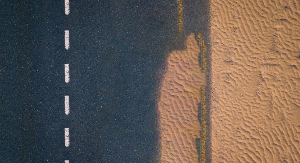 Luftaufnahme einer Straße mit gestrichelten weißen Linien auf dunklem Untergrund, rechts angrenzend an gewellten Sand. Die gerade Straße verläuft parallel zum strukturierten Sand und erzeugt einen Kontrast zwischen dem glatten Asphalt und der natürlichen Wüstenlandschaft – wie eine mit Photoshop perfektionierte Szene.