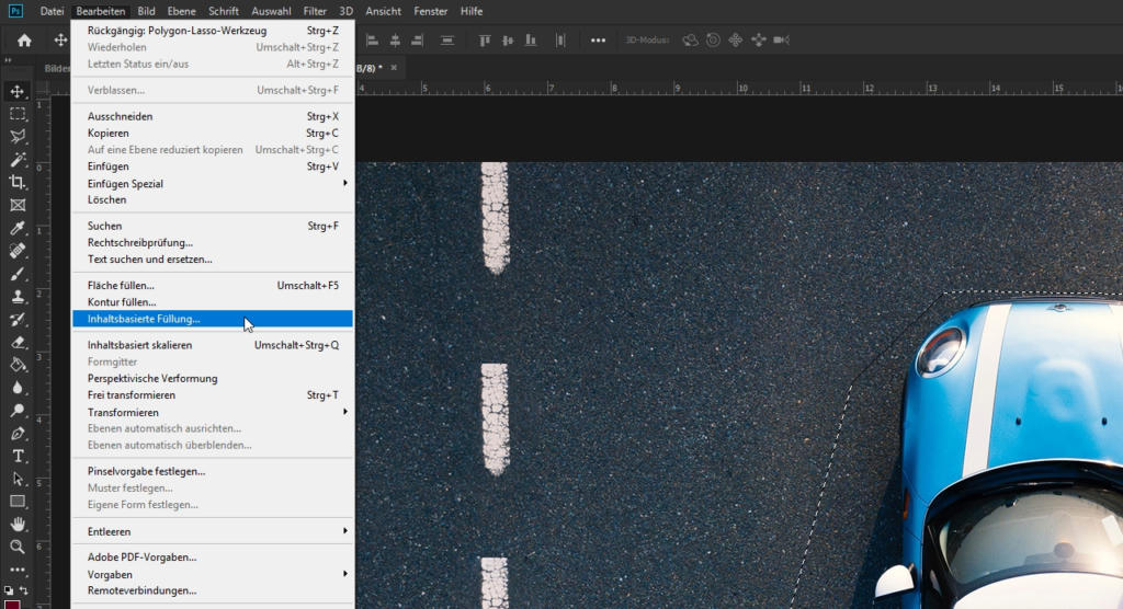 Ein teilweise sichtbares blaues Auto steht auf einer Straße mit gestrichelten weißen Linien. Oben links ist ein deutsches Optionsmenü eingeblendet, das Tools wie „Photoshop inhaltsbasiert füllen“ anzeigt. Dies lässt vermuten, dass es sich bei der Benutzeroberfläche um eine Bildbearbeitungssoftware handeln könnte.