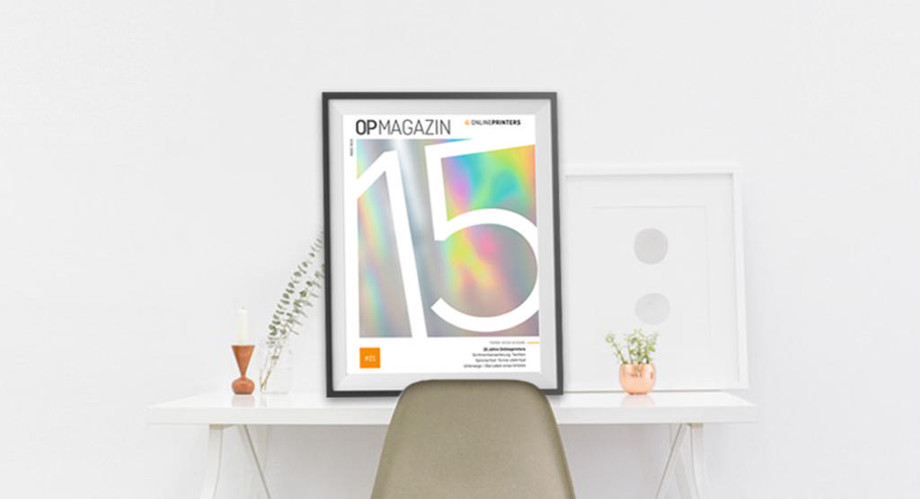 Ein minimalistischer weißer Schreibtisch mit einem gerahmten, abstrakten Cover der 15. Ausgabe einer Mitarbeiterzeitung lehnt an der Wand. Eine kleine Topfpflanze und eine Flasche mit einem einzelnen Stiel unterstreichen die Schlichtheit der Szene.