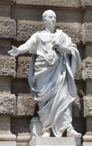 Statue einer gekleideten Figur, die mit erhobenem Arm gestikuliert und den anderen Arm an die Brust legt. Die Skulptur steht vor einer strukturierten Steinmauer. Auf dem Sockel steht „CICERONE“.