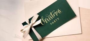 Eine grüne Karte mit dem Schriftzug „Christmas Party“ in eleganter Goldschrift und dem Titel „Einladung Weihnachtsfeier“ ist mit einem weißen Band zusammengebunden. Die Karte liegt auf einem beigen Umschlag vor einem weichen, unscharfen Hintergrund.