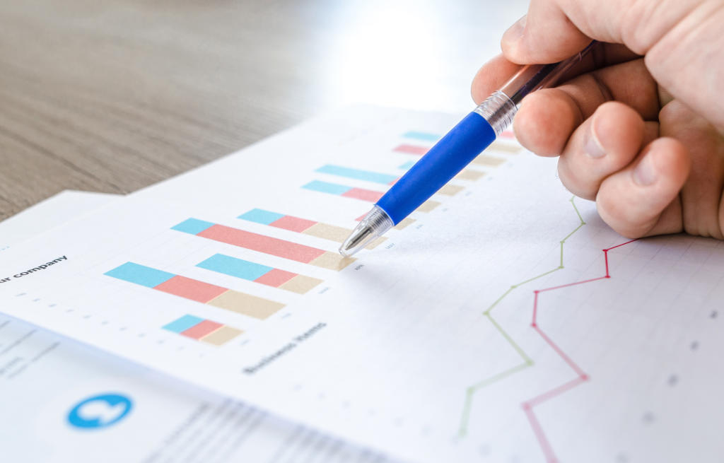 Eine Hand mit einem blauen Stift zeigt auf ein buntes Balken- und Liniendiagramm auf einem Blatt Papier, das Datenpunkte zum Beschwerdemanagement darstellt. Das Dokument liegt auf einer Holzoberfläche und zeigt Balken in Rot, Blau und Gelb sowie ein Liniendiagramm in Grün und Pink.