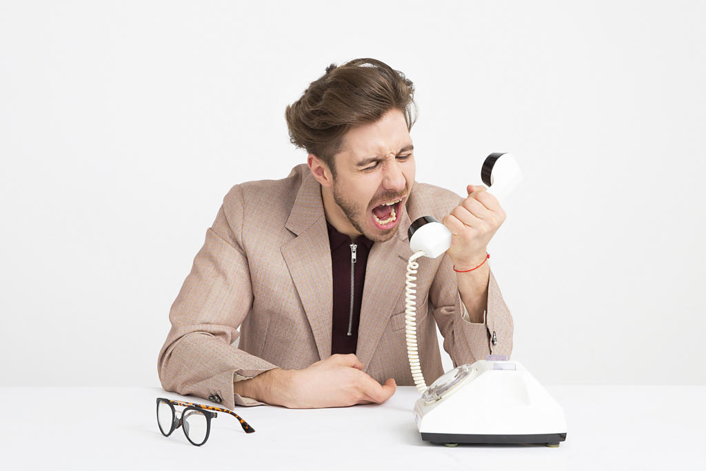 Ein Mann in beiger Jacke sitzt an einem weißen Tisch und vertieft sich in das Thema „Beschwerdemanagement“, während er in ein altmodisches weißes Telefon mit Wählscheibe brüllt. Sein Gesicht ist frustriert. Neben dem Telefon liegt eine Brille vor dem schlichten weißen Hintergrund.