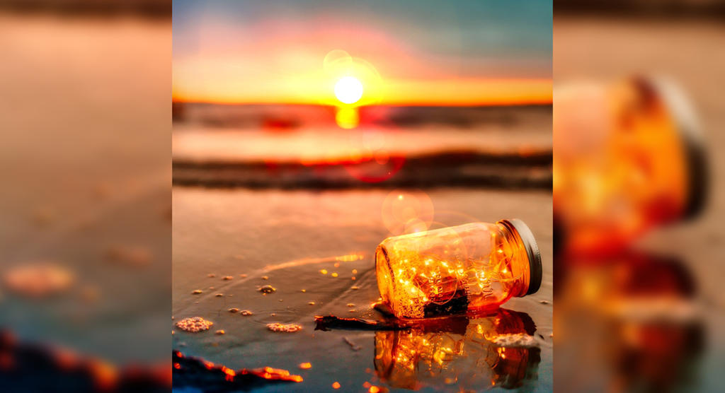 Ein Glas mit leuchtenden Lichterketten steht bei Sonnenuntergang an einem nassen Sandstrand. Die Sonne versinkt hinter dem Horizont und wirft ein warmes, goldenes Licht, das in seiner Perfektion fast wie Photoshop-gefiltert wirkt. Sanft plätschernde Wellen am Ufer spiegeln die leuchtenden Farben des Himmels wider.