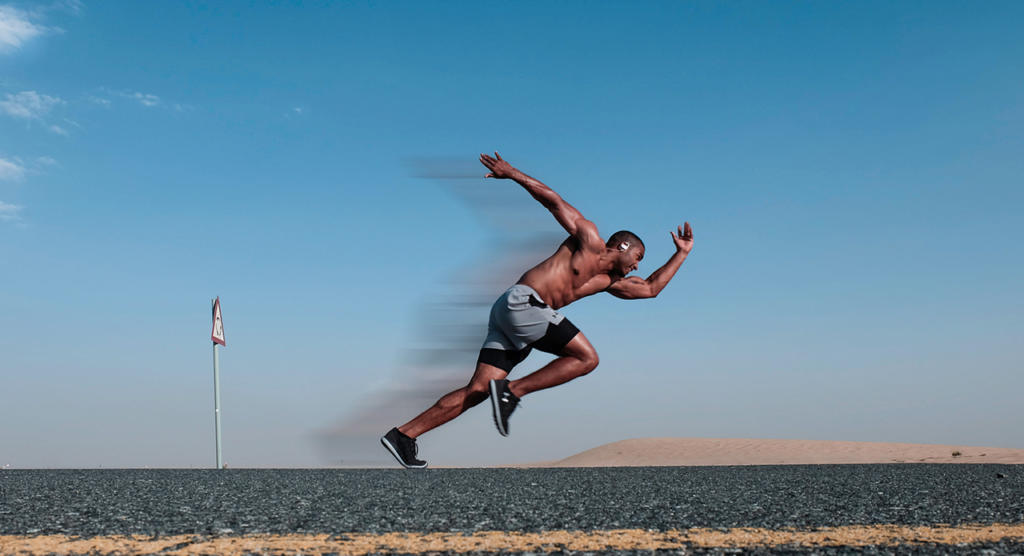 Ein Mann, dessen Geschwindigkeit durch einen Photoshop-Filter noch lebendiger wirkt, sprintet eine verlassene Straße in einer Wüstenlandschaft entlang. Oben ohne, in schwarzen Shorts und Turnschuhen, rast er unter einem klaren blauen Himmel an Sanddünen vorbei, wobei die Bewegungsunschärfe seine schnellen Bewegungen unterstreicht.