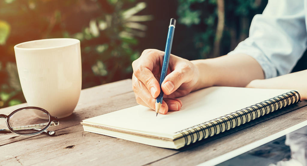 Ein Hauptredner macht sich an einem Holztisch im Freien mit einem blauen Stift Notizen in einem Notizbuch. Daneben stehen eine weiße Tasse und Gläser, während grünes Laub im Hintergrund verschwimmt und Sonnenlicht die ruhige Szenerie sanft erhellt.