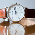 Eine minimalistische Armbanduhr, perfekt für einen Keynote-Speaker, mit weißem Zifferblatt, schwarzen Ziffern und Datumsfenster. Der Markenname „Rider“ ist elegant auf das Zifferblatt gedruckt. Das braune Lederarmband spiegelt sich wunderschön auf der glänzenden Oberfläche und verkörpert zeitlose Eleganz.
