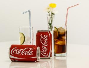 Zwei Coca-Cola-Dosen, eine stehend und eine liegend, neben zwei Gläsern. Ein Glas enthält Eis, eine Limettenscheibe und einen blauen Strohhalm. Das andere enthält Cola mit Limettenscheiben und einem rosa Strohhalm. Im Hintergrund steht eine Vase mit gelben und weißen Blumen.