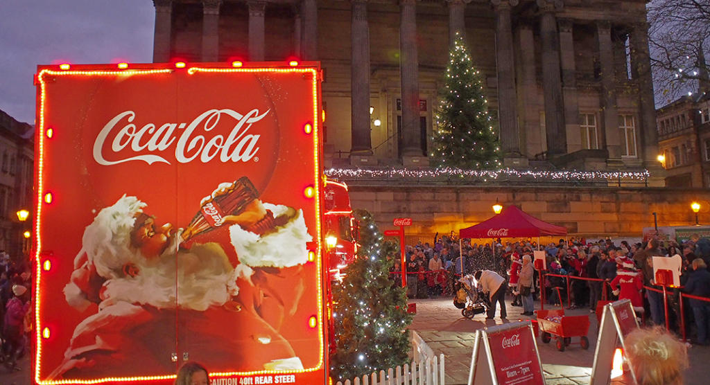 Eine festliche Szene entfaltet sich mit einem beleuchteten Coca-Cola-Truck, auf dem der Weihnachtsmann eine Cola genießt. In diesem perfekten Beispiel für Eventmarketing versammelt sich eine Menschenmenge um einen Weihnachtsbaum und ein festlich geschmücktes Gebäude, wobei die Weihnachtsbeleuchtung die fröhliche Atmosphäre verstärkt.