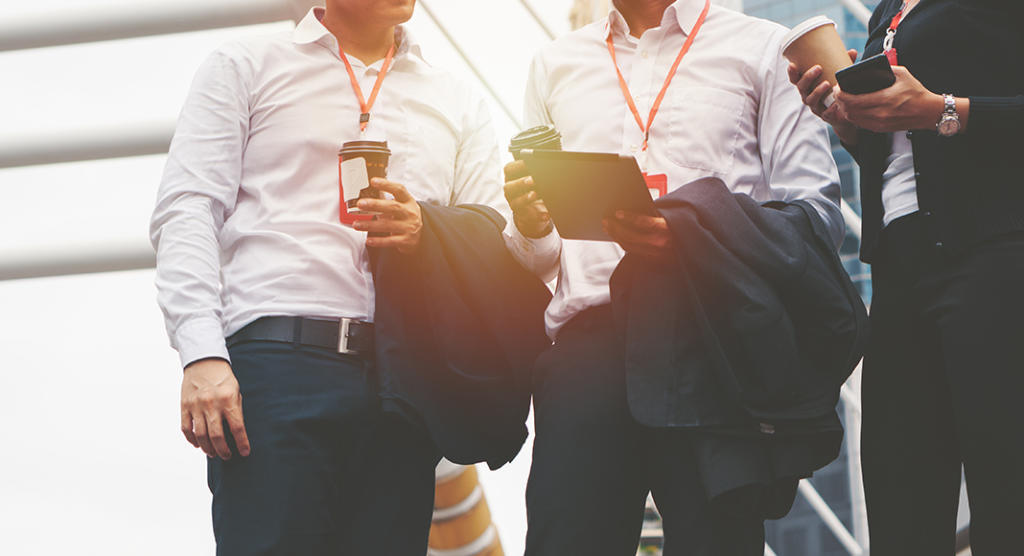 Drei Personen in weißen Hemden und mit Lanyards stehen im Freien und demonstrieren ihr Können im Eventmarketing. Zwei halten Kaffeetassen in der Hand, während eine andere mit über den Armen hängenden Jacken auf ihrem Smartphone hantiert. Eine helle Struktur verleiht der Szenerie Lebendigkeit, während eine Person mit einem Tablet arbeitet.