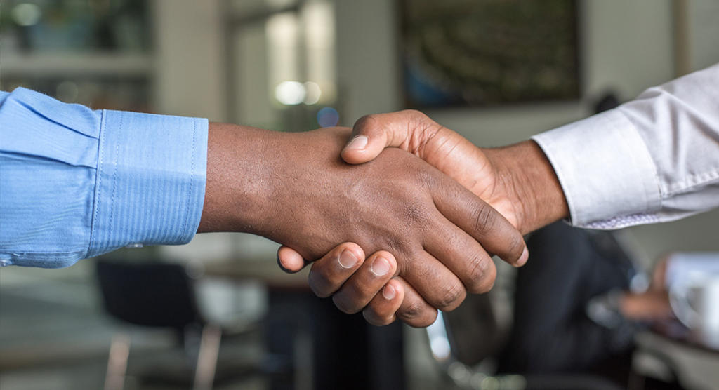 Zwei Personen geben sich in einem professionellen Umfeld die Hand und besprechen möglicherweise eine Event-Marketing-Strategie. Einer trägt ein blaues Hemd, der andere ein weißes. Büromöbel und ein unscharfer Hintergrund runden die Szene ab.