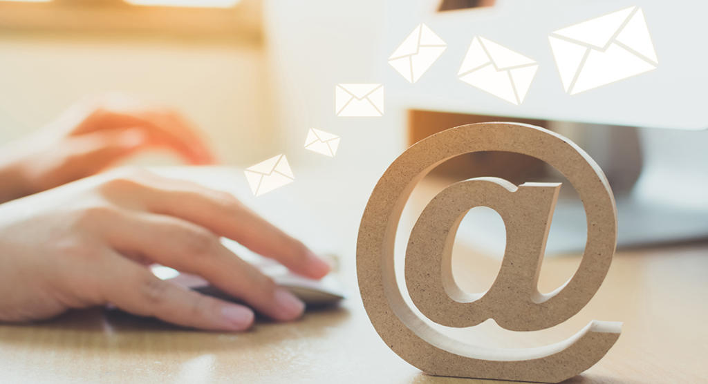 Eine Hand bedient eine Computermaus auf einem Schreibtisch, daneben ein hölzernes At-Zeichen. Darüber schweben mehrere Umschlagsymbole, die auf gesendete oder empfangene E-Mails hinweisen – möglicherweise Teil einer effektiven Event-Marketing-Strategie. Die Szene ist sanft von natürlichem Licht erhellt.