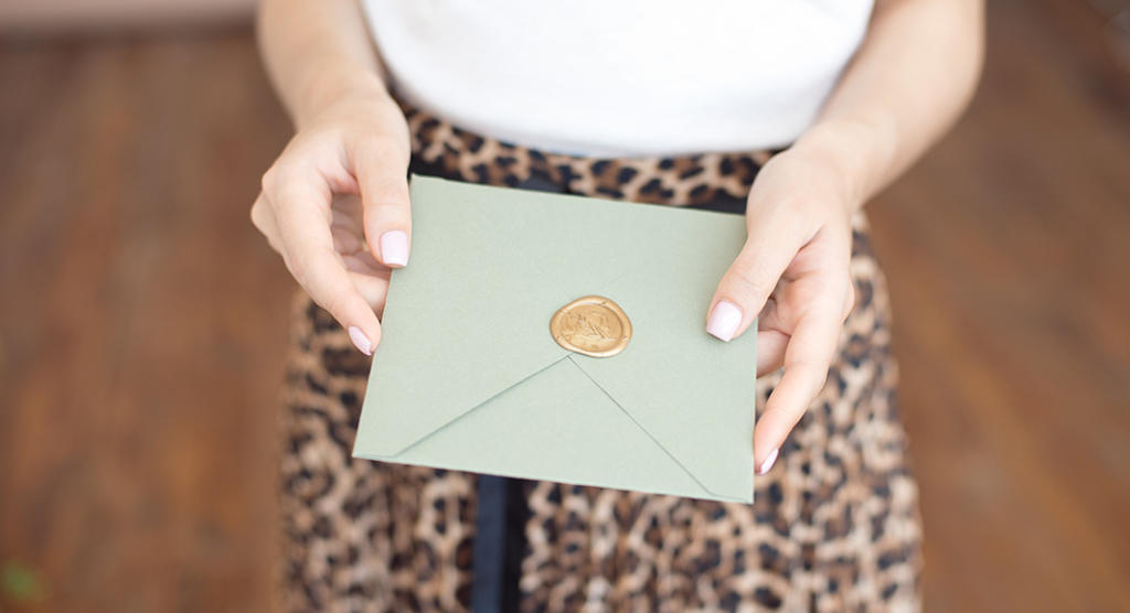 Eine Person hält einen grünen Umschlag mit einem goldenen Wachssiegel in der Hand, der auf eine exklusive Eventmarketing-Einladung hindeutet. Sie trägt einen Rock mit Leopardenmuster und ein weißes Oberteil, das sich vom unscharfen Hintergrund abhebt.