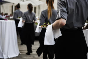 Kellner in grauen Hemden und schwarzen Schürzen tragen bei einer Veranstaltung im Freien Teller mit Salat. Sie laufen über eine Holzterrasse, an hohen, mit weißen Tischdecken bedeckten Tischen entlang, während im Hintergrund eine offene Atmosphäre herrscht.