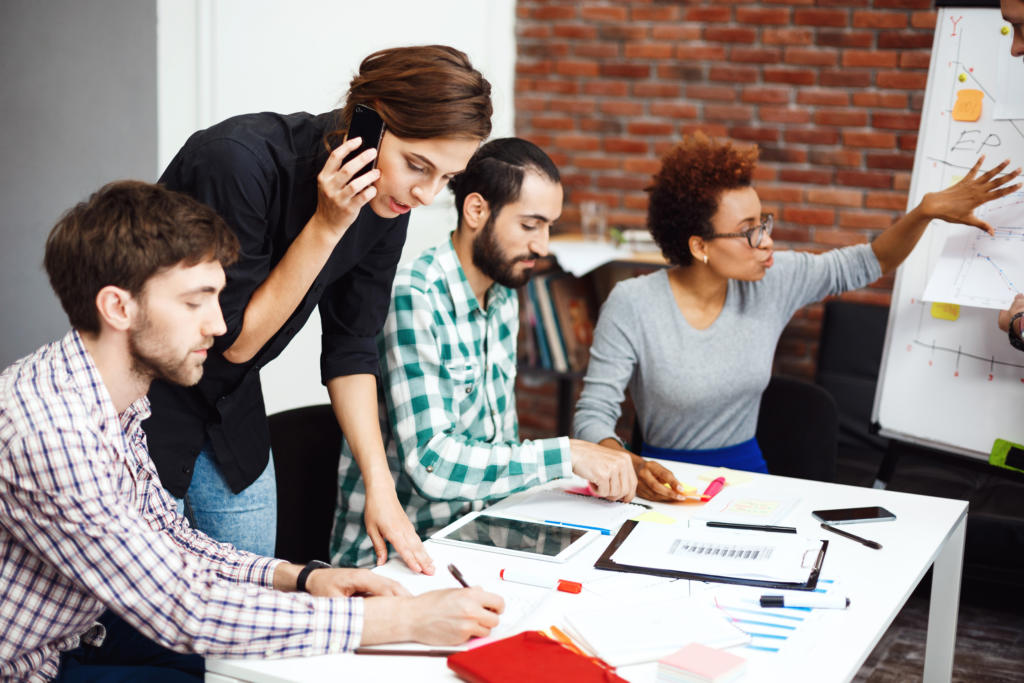 Vier Personen arbeiten in einem Büro zusammen, das an eine geschäftige Eventagentur erinnert. Einer telefoniert, während die anderen vor einer stilvollen Backsteinwand Dokumente und Diagramme prüfen. Ein Tablet und Papiere liegen auf dem Tisch und zeugen von organisierter Kreativität.
