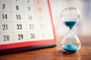 Links ist ein Kalender mit den Monatstagen zu sehen, rechts eine mit blauem Sand gefüllte Sanduhr. Die Sanduhr steht auf einer Holzfläche und symbolisiert den Lauf der Zeit.
