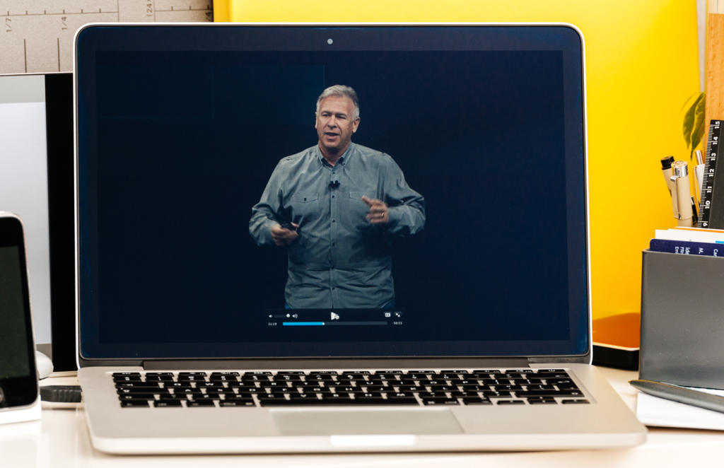 Auf einem Schreibtisch steht ein Laptop, auf dem ein Hauptredner steht und spricht. Um den Laptop herum befinden sich Bücher, ein Smartphone und Büromaterial. Eine gelbe Trennwand bildet den Hintergrund.