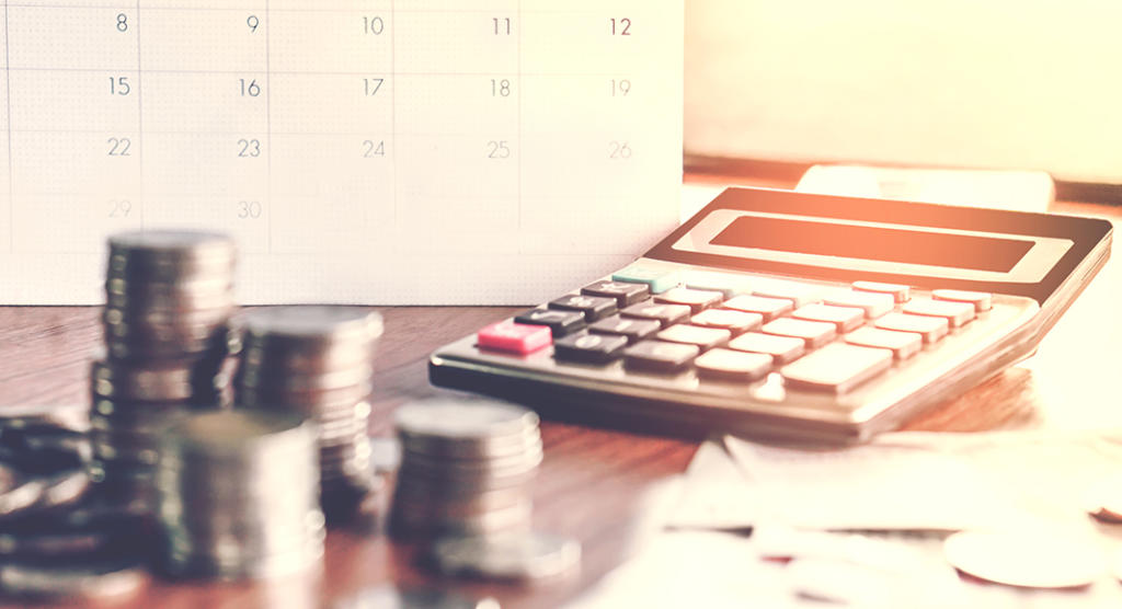 Münzstapel liegen auf einem Tisch neben einem Taschenrechner, im Hintergrund ist ein unscharfer Kalender zu sehen. In warmes Licht getaucht, erinnert diese Szene an die strategische Denkweise eines Hauptredners, der kluge Budgetierung und Finanzplanung betont.