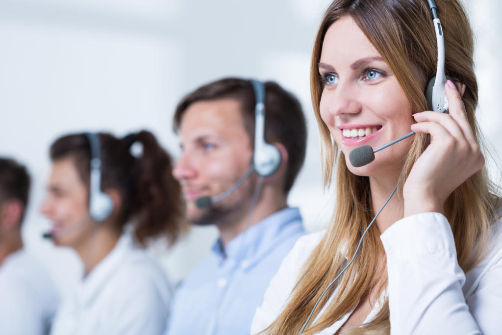 Eine Reihe von Callcenter-Agenten mit Headsets, die eifrig Kundenakquise betreiben, arbeiten an Computern. Im Vordergrund unterhält sich eine lächelnde Frau mit langen Haaren fröhlich. Im Hintergrund sind zwei weitere konzentrierte Personen in einer hellen und dynamischen Büroumgebung zu sehen.