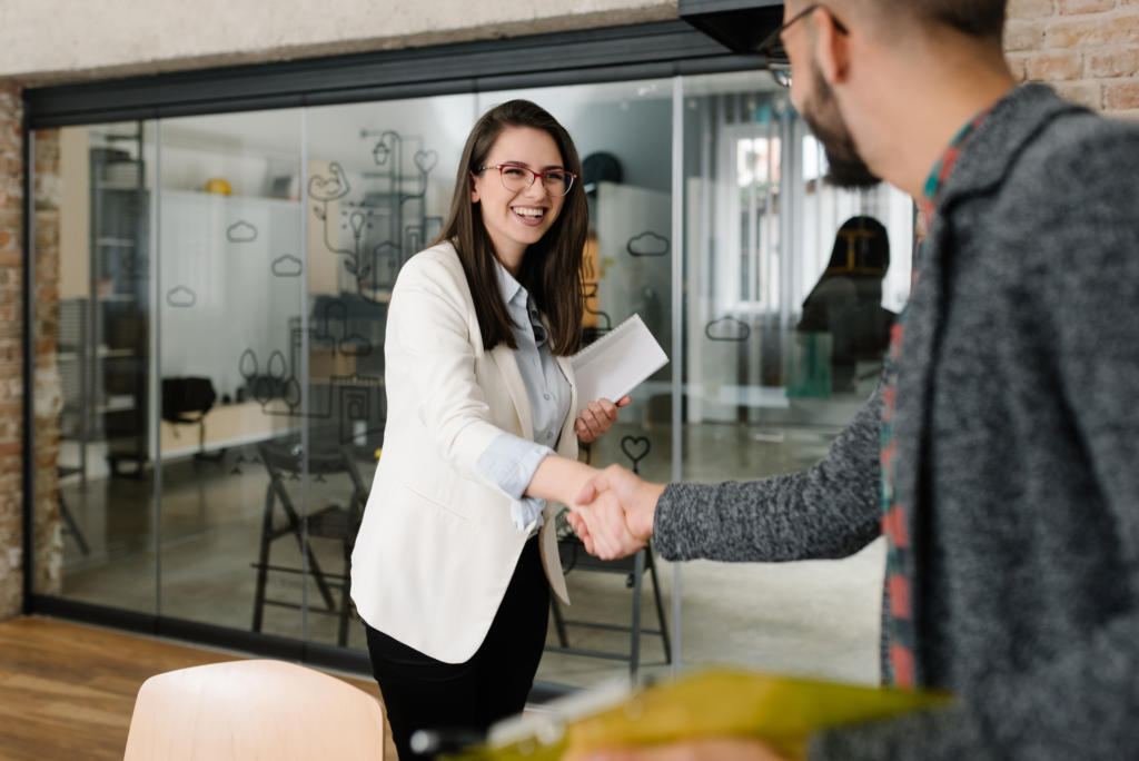 Zwei Personen befinden sich in einem Büro, das sich auf Kundenakquise konzentriert. Eine Person, mit Brille und weißem Blazer, lächelt und schüttelt einer anderen Person im grauen Pullover die Hand. Im Hintergrund ist ein verglaster Besprechungsraum zu sehen, der die freundliche Zusammenarbeit unterstreicht.