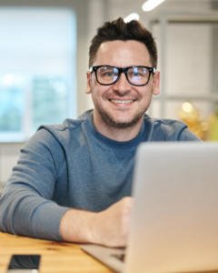 Eine Person mit Brille und blauem Pullover sitzt an einem Holztisch und lächelt in die Kamera. Sie nutzt einen Laptop, um die Kundenbindung zu stärken, daneben liegt ein Smartphone. Im Hintergrund ist ein heller Raum mit einem Fenster zu sehen.