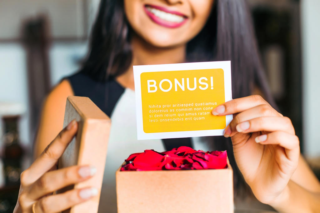 Eine Frau hält eine offene Schachtel mit roten Blütenblättern und einer Karte mit der Aufschrift „BONUS!“ im Rahmen einer Kundenbindungsaktion in der Hand. Sie lächelt und strahlt Freude und Freude aus. Der Hintergrund ist unscharf, wodurch die Aufmerksamkeit auf ihre Hände und die Schachtel gelenkt wird.