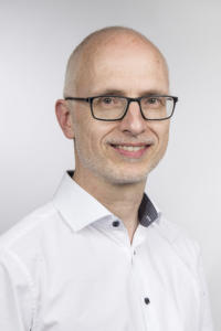 Ein lächelnder Mann mit Brille, Glatze und hellem Bart trägt ein weißes Hemd mit Kragen. Sein freundliches Auftreten spiegelt seine Expertise in der Kundenbindung wider. Der Hintergrund ist schlicht hellgrau.
