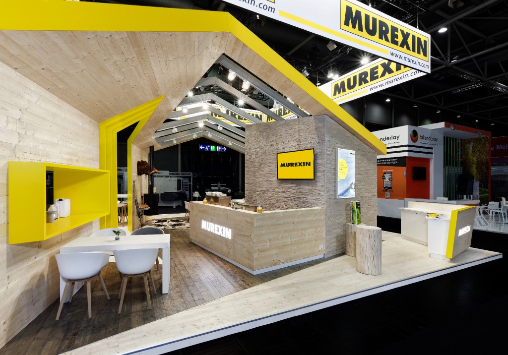 Erleben Sie innovatives Eventmarketing an unserem Messestand. Modern gestaltet mit Holzkonstruktion und leuchtend gelben Akzenten. Der Stand verfügt über einen Empfangstresen, einen Sitzbereich mit weißen Stühlen und eine markante Markenbeschilderung.