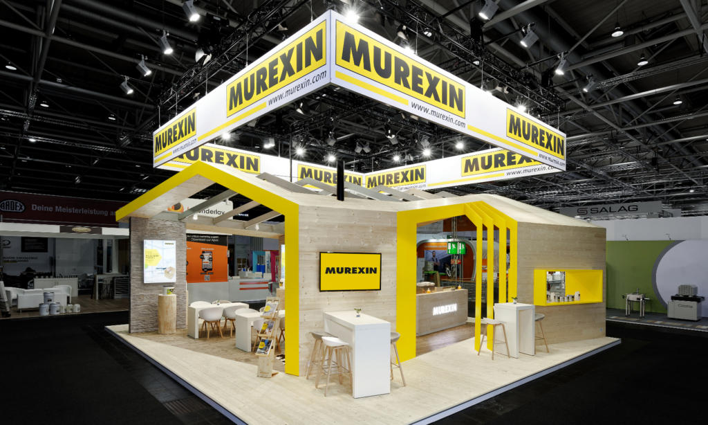Der Messestand von „Murexin“ präsentiert sich in modernem, kantigem Design mit leuchtend gelben Akzenten. Mit Sitzbereichen und Werbedisplays verkörpert der Raum stilvolles Eventmarketing. Ein Deckenschild präsentiert den Firmennamen prominent in der geräumigen Messehalle.