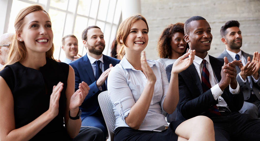 Eine bunt gemischte Gruppe von Männern und Frauen in Business-Kleidung sitzt in Reihen, lächelt und klatscht begeistert, während der Hauptredner seine Rede beendet. Die Atmosphäre erinnert an eine Konferenz oder ein Seminar mit großen Fenstern im Hintergrund.