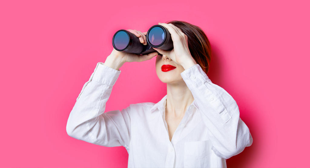 Eine Person mit leuchtend rotem Lippenstift und weißem Hemd, möglicherweise auf Talentsuche für eine Eventagentur, benutzt ein Fernglas vor einem leuchtend rosa Hintergrund. Sie blickt leicht nach oben, ihre kurzen Haare ergänzen die Szene perfekt.