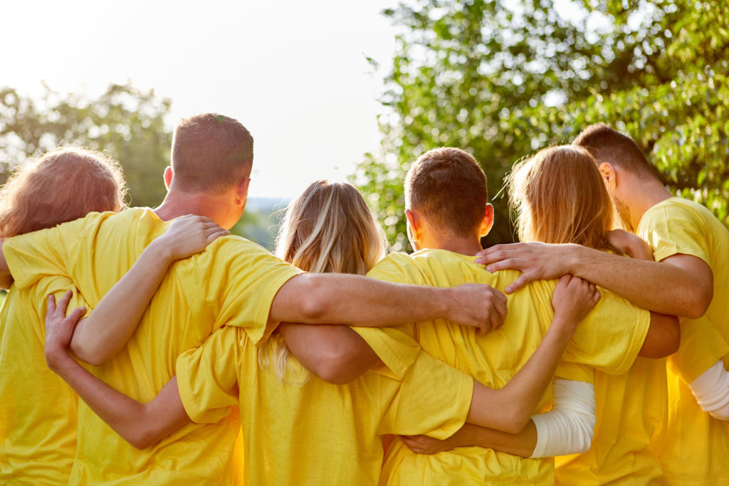 Eine Gruppe von Menschen steht im Freien mit dem Rücken zur Kamera und trägt passende gelbe Hemden. Sie umarmen sich mit den Armen um die Schultern, umgeben von grünen Bäumen und hellem Sonnenlicht.