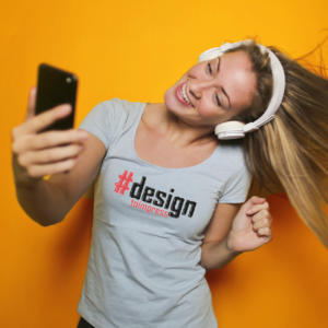 Eine lächelnde Person macht ein Selfie vor orangefarbenem Hintergrund. Sie trägt Kopfhörer und ein hellgraues Shirt mit der Aufschrift „#design to impress“. Die Haare fallen zur Seite.