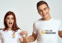 Zwei Personen tragen passende weiße T-Shirts mit dem Logo von „Online Printers“. Die linke Person zeigt auf ihr T-Shirt und lächelt mit offenem Mund, während die rechte Person breit lächelt und die Arme entspannt an den Seiten hängen lässt.