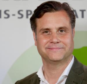 Ein Mann mit kurzen, dunklen Haaren lächelt leicht. Er trägt ein weißes Hemd mit Kragen und einen dunkelgrünen Blazer. Der Hintergrund ist verschwommen, Text und eine teilweise sichtbare grüne Form sind zu sehen.