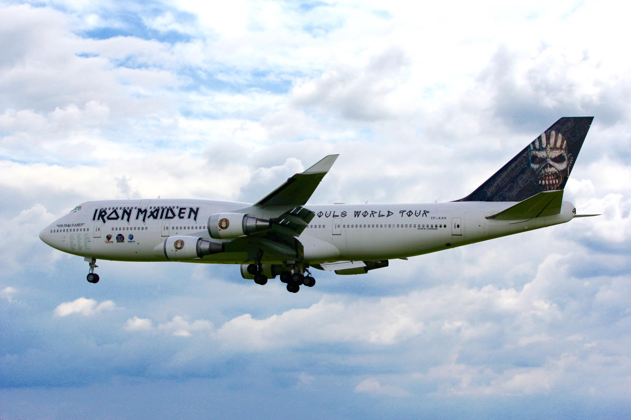 Eine weiße Boeing 747 im Flug, geschmückt mit dem legendären Iron Maiden-Schriftzug, trägt die Aufschriften „Iron Maiden“ und „World Tour“. Ein stilisierter Totenkopf ziert das Heck, während im Hintergrund Wolken den Himmel füllen.
