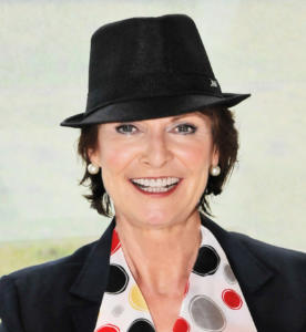 Eine lächelnde Person mit schwarzem Fedora-Hut, dunklem Blazer und buntem Schal mit kreisförmigen Mustern in Schwarz, Rot und Grau verkörpert die Essenz von Empfehlungsmarketing. Der helle, unscharfe Hintergrund verleiht diesem stilvollen Ensemble einen geheimnisvollen Touch.