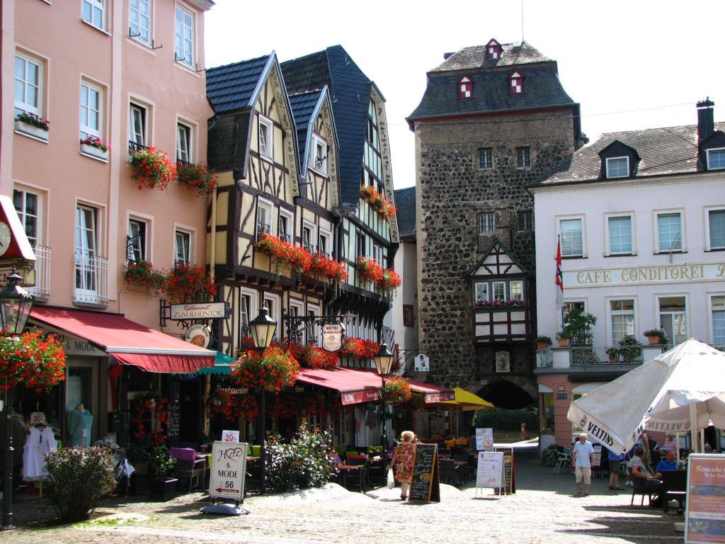 Eine charmante Straßenszene in einer europäischen Stadt mit farbenfrohen Fachwerkhäusern, geschmückt mit Blumenkästen. Im Hintergrund steht ein Steinturm, und Straßencafés mit Sonnenschirmen säumen die Kopfsteinpflasterstraße.