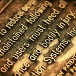 Nahaufnahme einer Metalltafel mit erhabenem englischen Text. Wörter wie „Benutzer“, „Achtung“ und „Körperverletzung“ sind sichtbar. Die Tafel hat ein gealtertes Bronze-Erscheinungsbild mit leicht unscharfem Hintergrund.