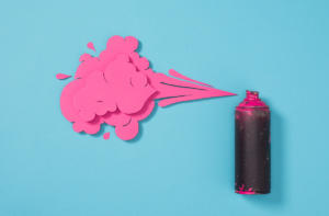 Eine Sprühdose sprüht eine Wolke aus rosa Papierausschnitten vor türkisfarbenem Hintergrund. Die Papierformen imitieren Sprühfarbe und erzeugen einen kreativen, künstlerischen Effekt.